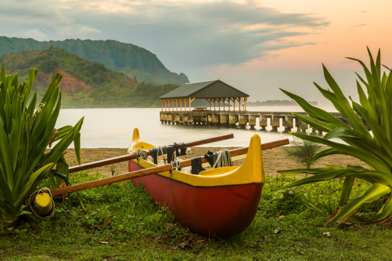 Red and yellow hawaiian canoe with outrigger on the beach at Hanalei pier at dawn as the sun lights the sky over Na Pali mountains