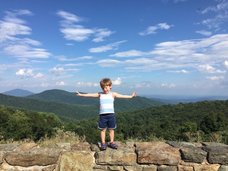 young boy standing on a rock wall along Skyline Drive in Shenandoah National Park with kids; background contains green rolling mountains and blue sky